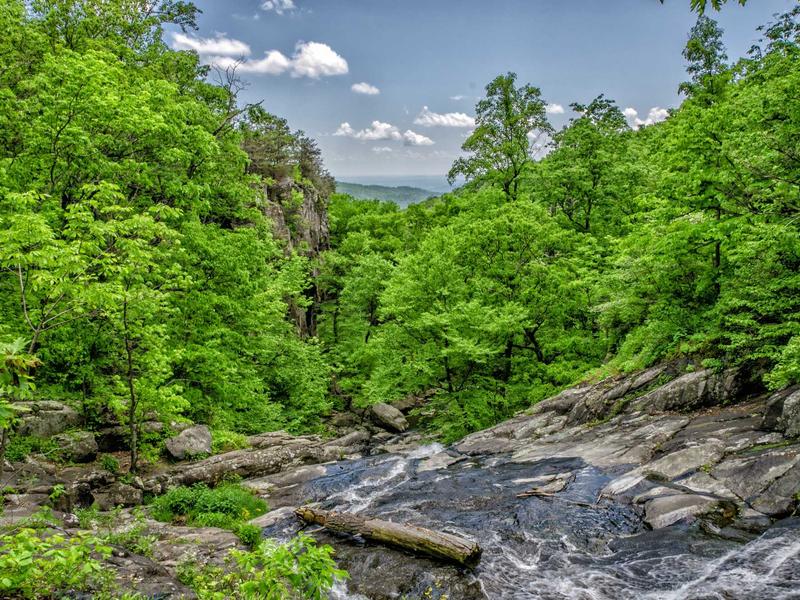 Waterfall flowing over gray rocks into a lush green forest valley with distant blue mountains